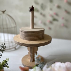 May include: A two-tiered wooden cake stand with a brown ribbon on top. The stand is made of natural wood and has a simple, elegant design.