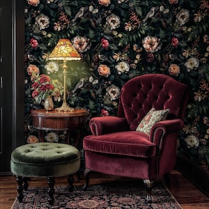 May include: A burgundy velvet armchair with a tufted back and a floral patterned pillow sits in front of a dark green wallpaper with a floral pattern. A green velvet ottoman sits in front of the chair. A stained glass lamp with a gold base sits on a wooden table next to the chair.