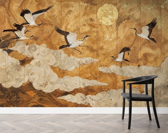 Puede incluir: Un mural con grullas en vuelo sobre un fondo dorado y texturizado con un gran sol circular. Las grullas tienen alas negras y blancas y marcas rojas. Las nubes son de color beige claro. Una silla negra está en primer plano.