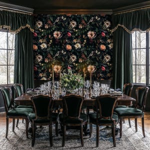 May include: A dark wood dining table set with black velvet chairs. The table is set with a floral centerpiece and glassware. The room has dark green velvet curtains and a floral wallpaper.