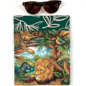 May include: A rectangular fabric sleeve featuring a vibrant jungle print with a lion, leopard, zebra, and tropical bird, topped with a dark green section with a white leaf pattern. A pair of brown tortoiseshell sunglasses rests above the sleeve.