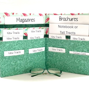 May include: A fabric organizer with multiple pockets featuring a light green floral pattern and a top border with a red strawberry print. The pockets contain white cards labeled Magazines, Brochures, Notebook or Tall Tracts, New Tracts, and Contact Cards.