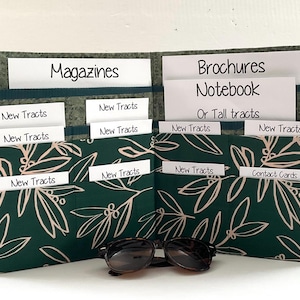 May include: A dark green fabric organizer with a light-colored leaf pattern, featuring multiple pockets labeled with white cards reading Magazines, Brochures, Notebook, Or Tall tracts, New Tracts, and Contact Cards, with sunglasses in front.