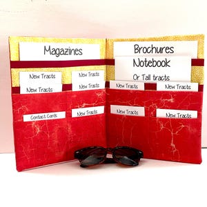 May include: Open organizer with red textured pockets and yellow patterned accents. Labeled "Magazines" and "Brochures Notebook Or Tall tracts." Includes pockets for "New Tracts" and "Contact Cards." Sunglasses rest in front.