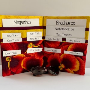 May include: A fabric organizer with a yellow and red floral pattern featuring multiple pockets labeled Magazines, Brochures, Notebook or Tall Tracts, New Tracts, and Contact Cards. A pair of dark-rimmed sunglasses rests in front of the open organizer.