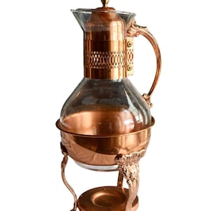 May include: A copper-colored glass carafe with a decorative handle and lid. The carafe sits on a matching copper stand with ornate legs and a round base. The glass portion of the carafe is clear, allowing the contents to be visible.