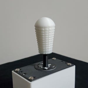 May include: White plastic gear shift knob with a textured surface. The knob is attached to a black metal shaft and is mounted on a black metal base.