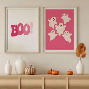 May include: Two framed prints on a light beige wall. The print on the left says "BOO!" in pink letters. The print on the right has a pink background with five white ghosts with black eyes.