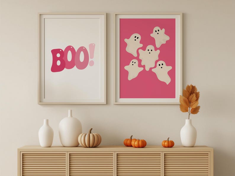May include: Two framed prints on a light beige wall. The print on the left says "BOO!" in pink letters. The print on the right has a pink background with five white ghosts with black eyes.