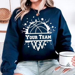May include: Navy blue sweatshirt with a white basketball graphic and the text "YOUR TEAM" above a basketball hoop. The graphic has white paint splatters around it.