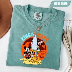 May include: Light green t-shirt featuring Mickey Mouse, Pluto, Goofy, and Donald Duck with a surfboard. The shirt has the text "King of the Wave" and "Making Waves since 1928" in orange and red. A silver necklace with a pendant is on the shirt.
