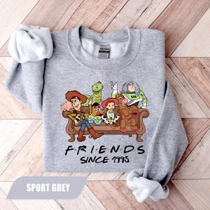 May include: Sport grey sweatshirt featuring a graphic of Toy Story characters on a brown couch. The text "FRIENDS SINCE 1995" is printed below the characters. The sweatshirt is made of a soft material.