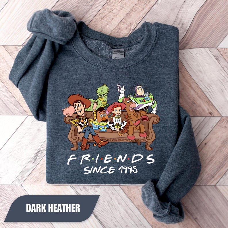 May include: Dark heather gray sweatshirt featuring a graphic of Toy Story characters on a brown couch. The text "FRIENDS SINCE 1995" is below the graphic. The bottom left corner of the sweatshirt has a dark gray rectangle with the words "DARK HEATHER".