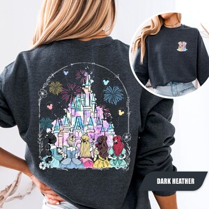 May include: Dark heather gray sweatshirt featuring a colorful castle graphic with fireworks and Disney princess silhouettes. The design includes floral and Mickey Mouse details. The sweatshirt has a crew neck and long sleeves. The text "DARK HEATHER" is at the bottom right.