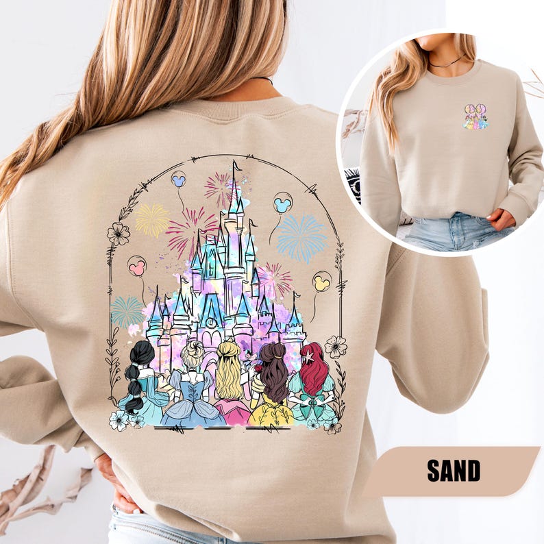 May include: Sand-colored sweatshirt featuring a colorful graphic of a castle with fireworks and several figures. The design is framed by a floral arch. A smaller image on the right shows a close-up of the upper chest with a colorful design. The word "SAND" is at the bottom.