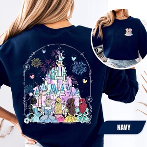 May include: Navy blue sweatshirt featuring a colorful illustration of a fairytale castle with fireworks and Disney princesses. The design is framed by a floral arch and Mickey Mouse balloons. A smaller image of the design is on the upper right.