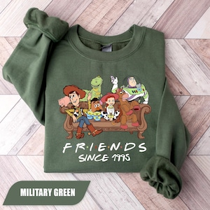 May include: Military green sweatshirt featuring a graphic of characters from the animated film Toy Story. The design includes Woody, Buzz Lightyear, and other characters on a brown couch. The text "FRIENDS SINCE 1995" is printed below the graphic.