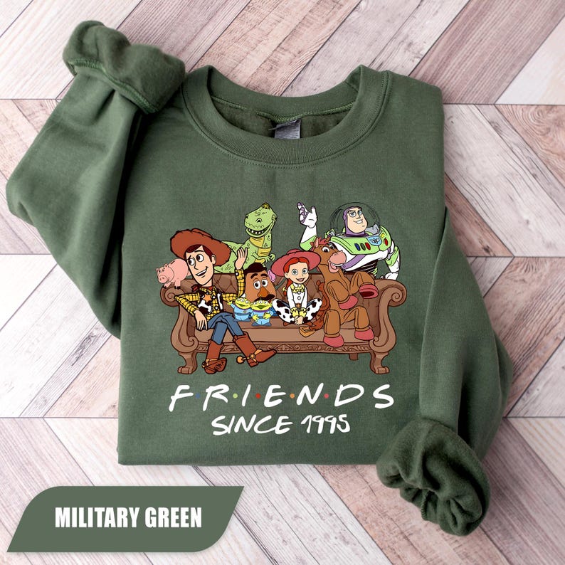 May include: Military green sweatshirt featuring a graphic of characters from the animated film Toy Story. The design includes Woody, Buzz Lightyear, and other characters on a brown couch. The text "FRIENDS SINCE 1995" is printed below the graphic.