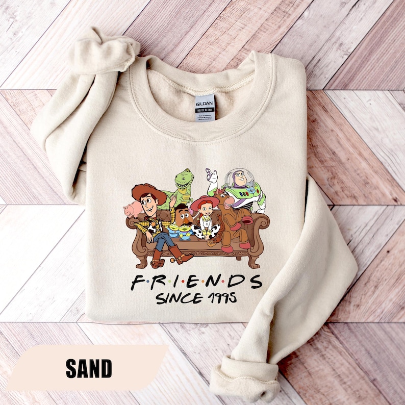 May include: Sand-colored sweatshirt featuring a graphic of Toy Story characters on a brown couch. The text "FRIENDS SINCE 1995" is below the image. The sweatshirt has a crew neck and long sleeves.