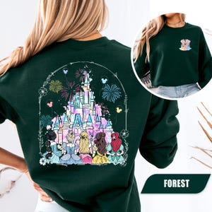 May include: A dark green sweatshirt featuring a colorful illustration of a castle with fireworks and several princess figures. The design is framed by a white arch with floral and balloon accents. The word "FOREST" is printed at the bottom.
