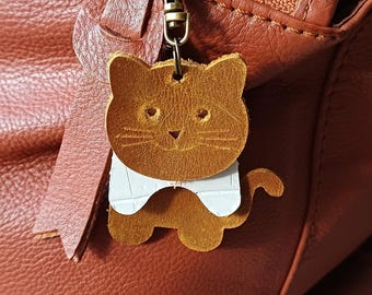Op de afbeelding: Een bruinleren katvormige sleutelhanger met een witte kraag, bevestigd aan een koperen ritstrekker op een roodbruine leren tas. De hanger heeft gestikte details voor de ogen, neus en snorharen.