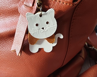 Op de afbeelding: Een witte katvormige leren bedel met een bruine vlinderdas hangt aan een bronskleurige sluiting die aan een bruine leren tas is bevestigd. De tas heeft een rits en een riem. De bedel is een decoratief accessoire.