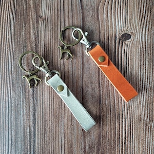 Puede incluir: Dos llaveros de cuero con herrajes metálicos y dijes de perro. Un llavero es plateado y el otro naranja. Ambos llaveros tienen un cierre a presión y un anillo metálico para llaves.