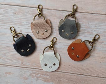 Op de afbeelding: Vijf leren sleutelhangers in de vorm van kattengezichten in verschillende kleuren: zwart, grijs, beige, wit en bruin. Elke sleutelhanger heeft een gouden metalen ring en een kleine metalen lus.