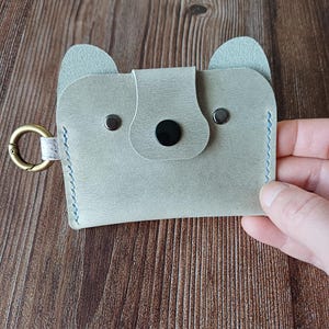 Può includere: Portacarte in pelle grigio chiaro a forma di muso di orso, con bordi cuciti blu e un naso a bottone nero. Presenta due piccole orecchie e un anello metallico color oro per attaccare chiavi o un cordino.