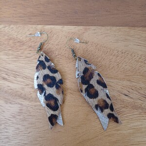 Könnte beinhalten: Ein Paar Ohrhänger mit Leopardenmuster. Die Ohrringe haben eine Federform mit einem braunen und schwarzen Muster und einem silbernen Glitzerakzent am unteren Ende. Sie haben goldfarbene Ohrhaken. Ein modisches Accessoire.