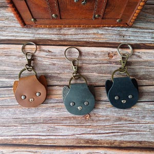 May include: Three leather cat-shaped keychains in brown, gray, and black. Each keychain has a metal ring and clasp. The keychains are on a wooden surface, with a brown leather chest in the background.