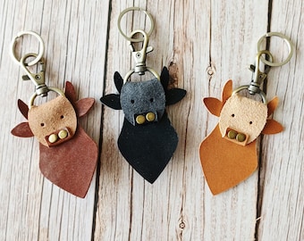 Pode incluir: Três porta-chaves de couro em forma de cabeça de vaca, cada um com um mosquetão e um anel de metal. O da esquerda é castanho escuro, o do centro é preto e o da direita é castanho claro. Cada um tem um focinho mais claro com dois rebites.