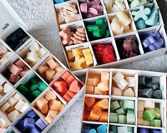 May include: Assortment of colorful wax melts in various shapes and scents, arranged in individual compartments within white boxes. The melts include heart, star, and geometric shapes in shades of blue, green, pink, orange, and yellow. The boxes are labeled with scent names.