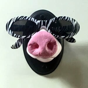 May include: A black and white zebra-print sunglasses with a pink pig nose attached to a black circular base.