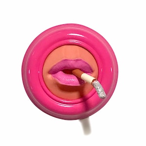 Peut inclure: Un cendrier en céramique rose en forme de bouche avec une cigarette qui en sort. La bouche est peinte avec du rouge à lèvres rose et la cigarette est blanche avec un filtre gris.