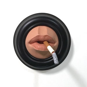 May include: A close-up of a sculpted mouth with a cigarette in it. The mouth is painted a light brown color and the cigarette is white with a brown filter. The mouth is framed in a black circular frame.