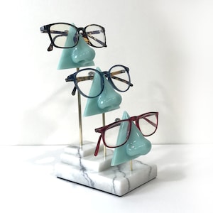 May include: A white marble display stand with three tiers, each featuring a light blue nose-shaped stand with a pair of glasses resting on it. The glasses are tortoise shell, blue, and red.