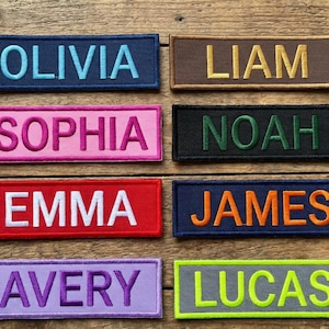 Może przedstawiać: Prostokątne naszywki z haftowanymi imionami: Olivia, Liam, Sophia, Noah, Emma, James, Avery i Lucas. Każda naszywka ma inną kombinację kolorów, z imionami w kontrastowych kolorach nici.