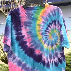 Könnte beinhalten: Ein kurzärmeliges T-Shirt mit Batikmuster und Spiraldesign. Das Shirt zeigt eine lebendige Mischung aus Farben, darunter Blau, Rosa, Lila, Gelb und Grün. Das Shirt hängt an einem schwarzen Kleiderbügel.