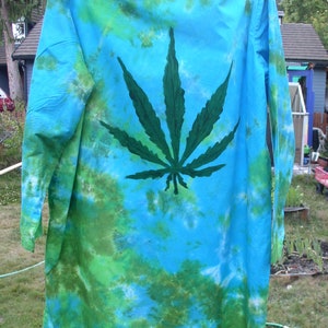 May include: A long-sleeved shirt with a tie-dye pattern in shades of blue and green. A large, dark green cannabis leaf is prominently displayed on the back. The shirt is hanging on a clothesline outdoors.