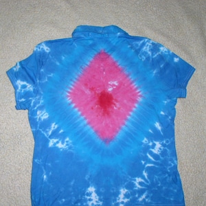 May include: Blue tie-dye polo shirt with a large pink diamond design on the back. The shirt has short sleeves and a collared neckline. The tie-dye pattern includes white accents.