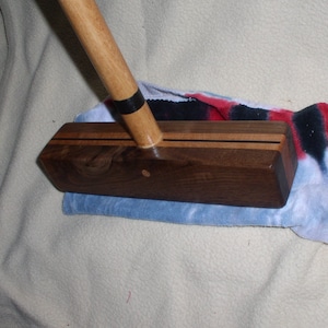 May include: A wooden croquet mallet with a dark brown head and a light brown handle. The head has two thin black stripes and is resting on a blue and red towel. The handle is attached to the head with a black band.