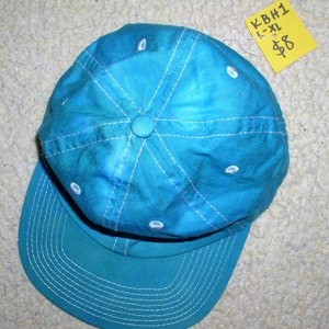 May include: A turquoise baseball cap with a curved bill. White stitching accents the panels and bill. The cap has a button on top and small white eyelets. A yellow tag with the text "KBH1 L-XL" is attached.