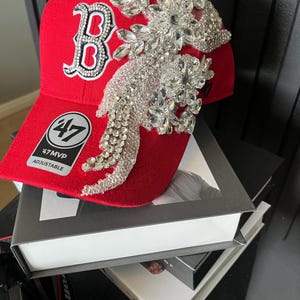 May include: A red baseball cap with a white and crystal-studded "B" logo. The cap features a large, ornate crystal embellishment on the side and the text "47 MVP ADJUSTABLE". The cap is sitting on top of a stack of books.