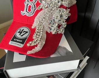 Könnte beinhalten: Rote Baseballkappe mit weißem und kristallbesetztem "B"-Logo. Die Kappe hat eine große, verzierte Kristallverzierung an der Seite und den Text "47 MVP ADJUSTABLE". Die Kappe liegt auf einem Stapel Bücher.