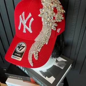 Op de afbeelding: Rode baseballcap met een wit geborduurd "NY" logo en een sprankelende kristallen versiering. De cap heeft een gebogen klep en een verstelbare band. De cap heeft een "47 MVP" patch.