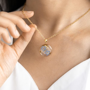 May include: A gold chain necklace with a round pendant featuring a delicate arrangement of blue forget-me-not flowers pressed inside clear resin.