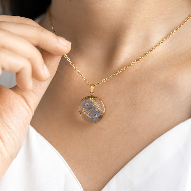 May include: A gold chain necklace with a round pendant featuring a pressed flower design. The pendant is filled with blue and white flowers.