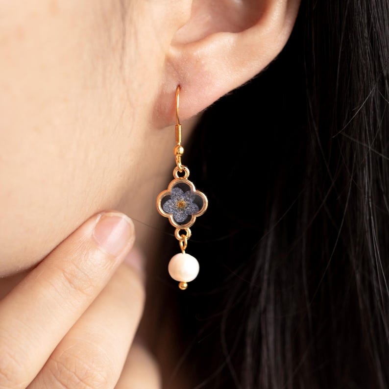 May include: A gold-coloured earring with a four-leaf clover design, featuring a blue and yellow floral pattern, and a dangling white pearl. The earring is worn on a person's ear.
