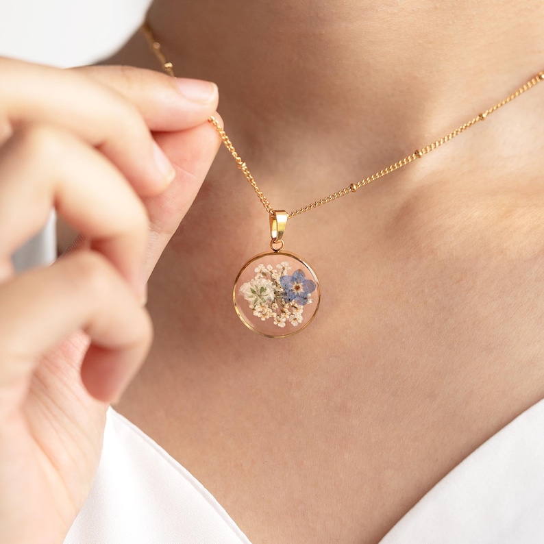 May include: A gold chain necklace with a round pendant featuring pressed flowers in shades of blue and white.